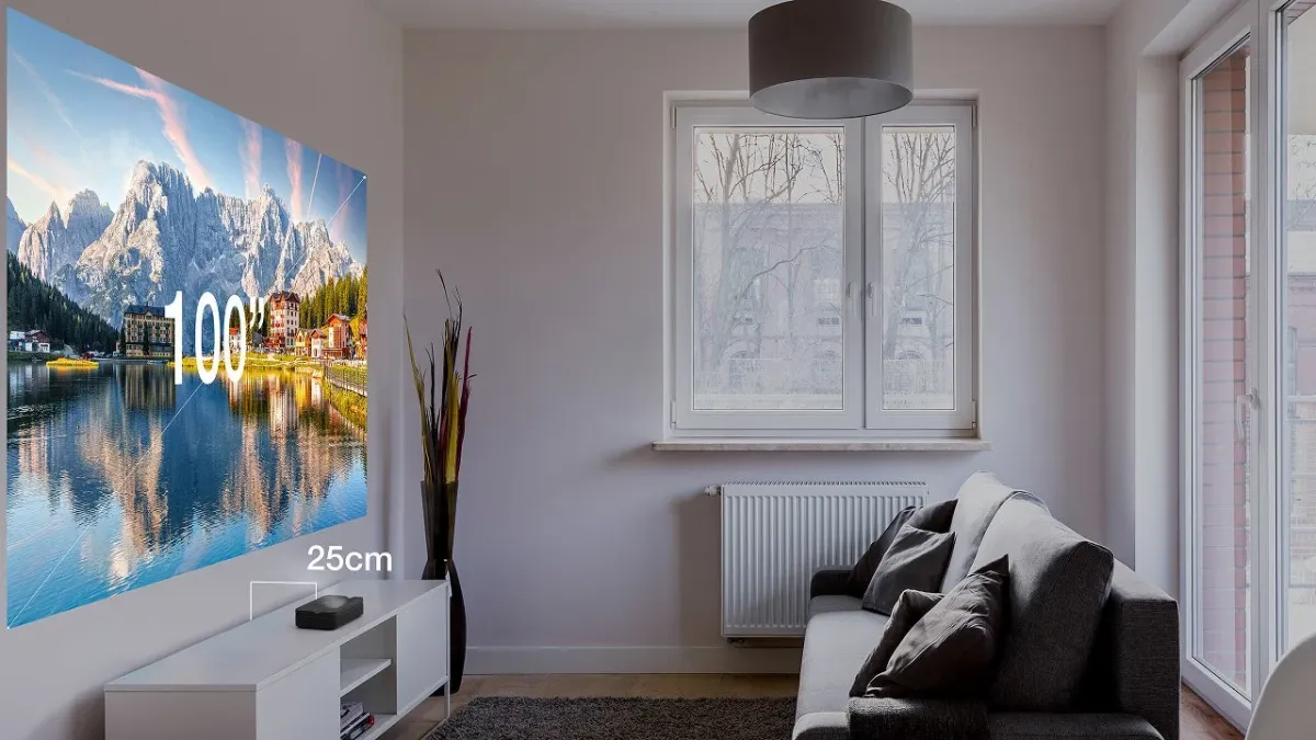 リビングルームで超短焦点プロジェクターが壁に100インチの美しい湖と山々の景色を投影しています。プロジェクターは壁からわずか25cmの距離に置かれており、省スペースで大画面を楽しめる様子が分かります。