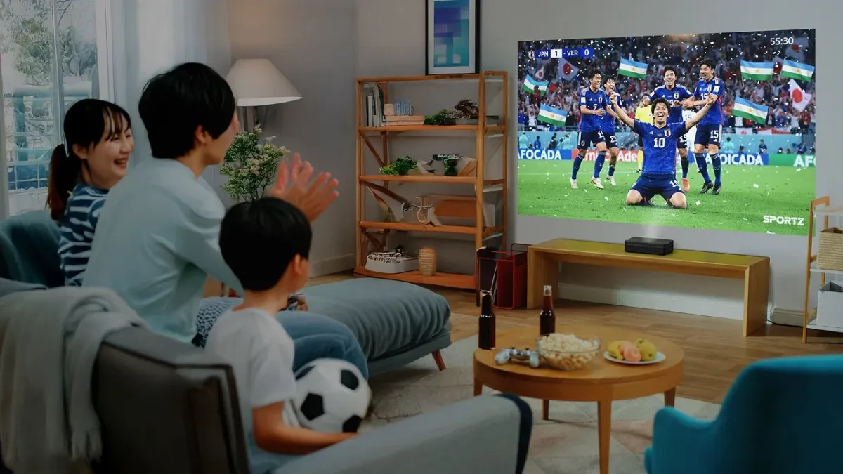 リビングで家族がプロジェクターを使い、サッカー日本代表の試合を観戦している。ゴールが決まり、壁に映し出された選手たちの喜びの瞬間を拍手しながら、ポップコーンと共に楽しんでいる様子。