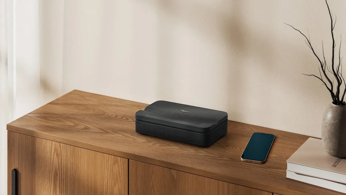 木製のキャビネットの上に、黒いプロジェクター、スマートフォン、そして枝物の入った花瓶が置かれた、ミニマリストでモダンなインテリアの部屋の様子。落ち着いた色合いで統一されています。