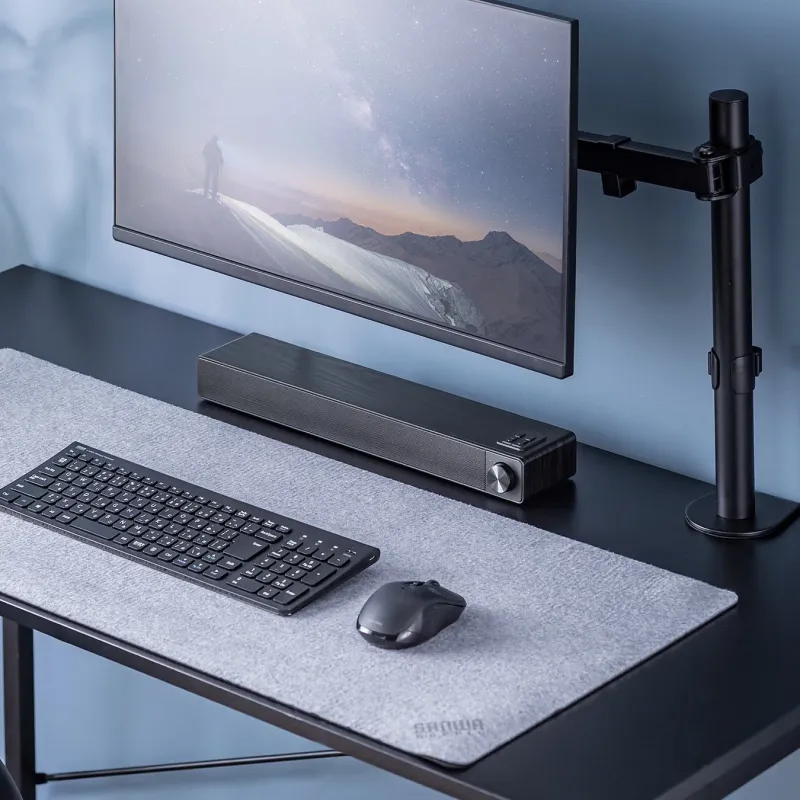 モダンなPCデスクのセットアップが写っています。モニターには雪山の風景が映し出され、その手前にはキーボード、マウス、サウンドバーが配置されています。シンプルで機能的なワークスペースです。