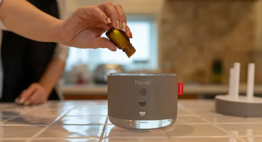 女性がグレーのアロマディフューザーに小さなボトルからエッセンシャルオイルを注いでいる様子です