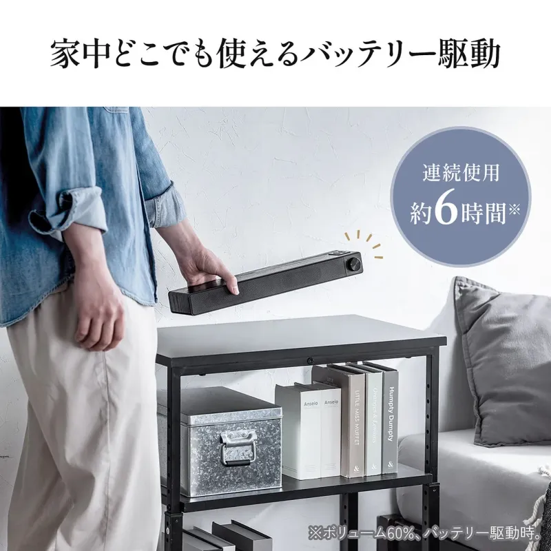 家中どこでも使えるバッテリー駆動のサウンドバーを持つ人物の画像です。連続使用約6時間（ボリューム60%時）が可能で、リビングの棚に置かれた本や小物と共に、快適なホームエンターテイメントを提案しています。