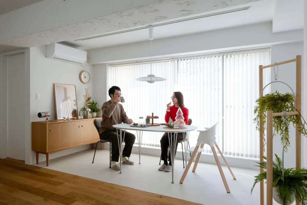 明るくモダンなリビングダイニングで、男女のカップルがテーブルを囲み、リラックスしてコーヒータイムを楽しんでいる様子です。観葉植物や木製家具が配置され、落ち着いた雰囲気が漂っています。