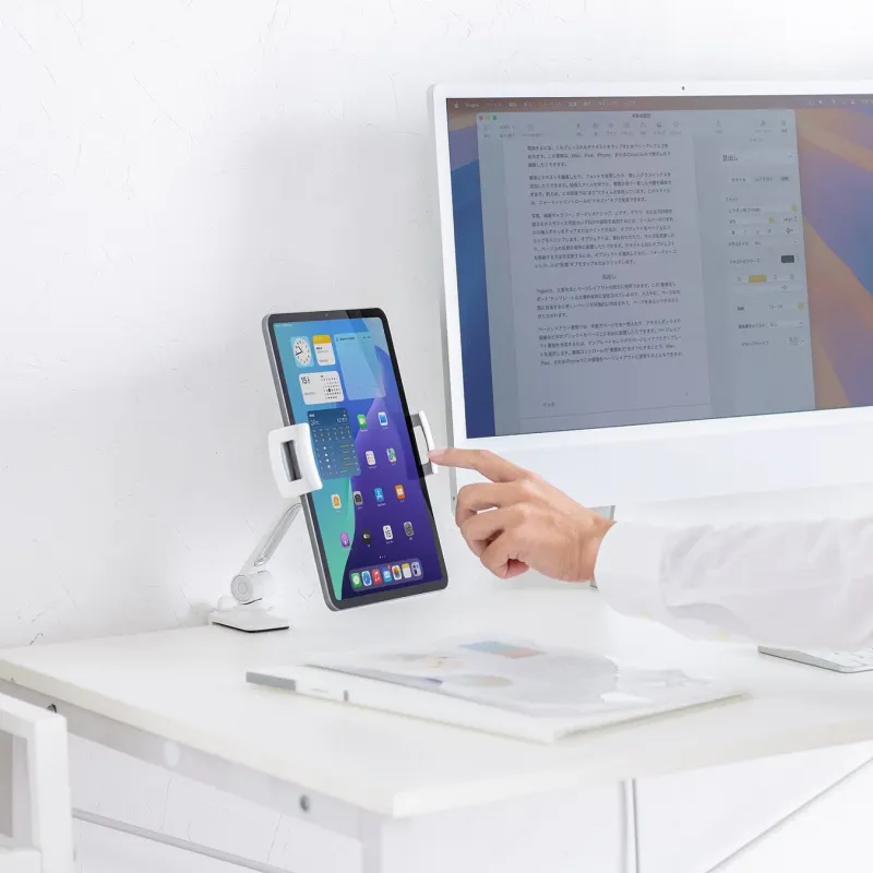 白いデスク上に、調整可能なスタンドに置かれたiPadと大型モニターが並び、手でタブレットを操作している様子。モニターには文書編集画面、タブレットにはホーム画面が表示されており、効率的な作業環境がうかがえます。