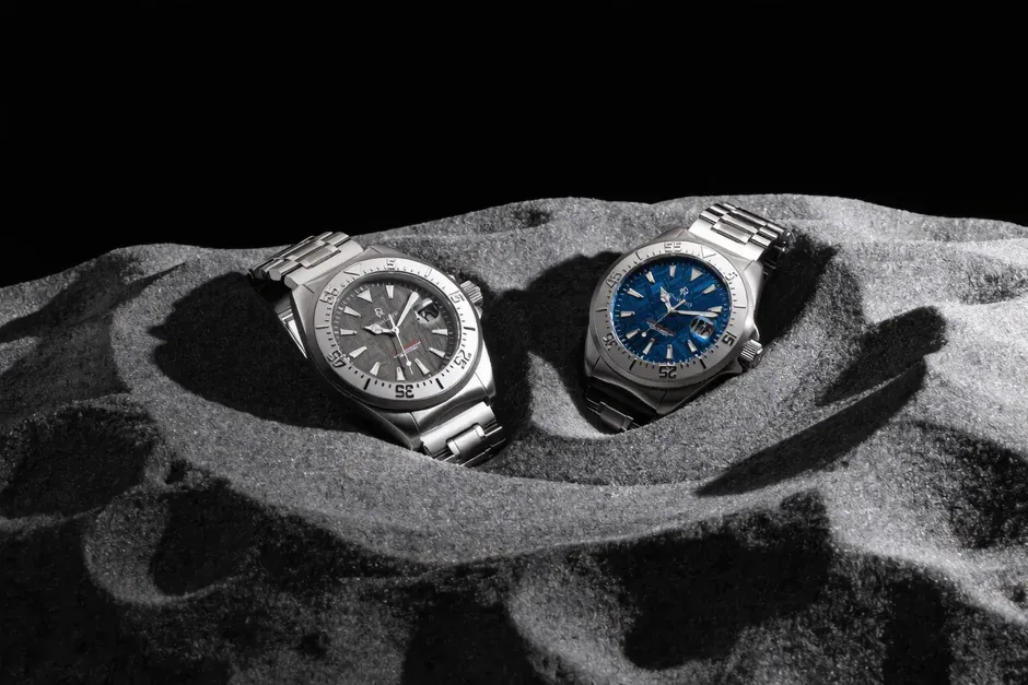 月面を思わせる灰色の砂地の上に置かれた文字盤がグレーとブルーの2つの金属製腕時計