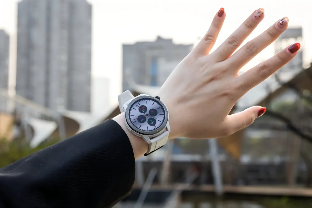 白いスマートウォッチを腕に着けた人物の手元が写っています。背景には都市の建物がぼやけて見え、スマートウォッチの画面にはデータが表示されています。指にはおしゃれなネイルが施されており、現代的なライフスタイルを想起させます。