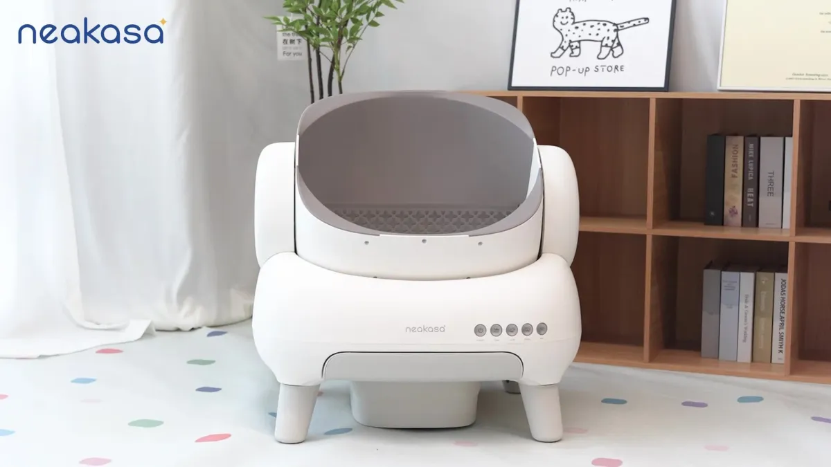 白い「neakasa」ブランドの自動猫用トイレがモダンな部屋の中央に置かれている。背景には本棚、植物、猫のイラストがあり、カラフルな水玉模様の床が特徴的。スマートなペット用品の設置例。