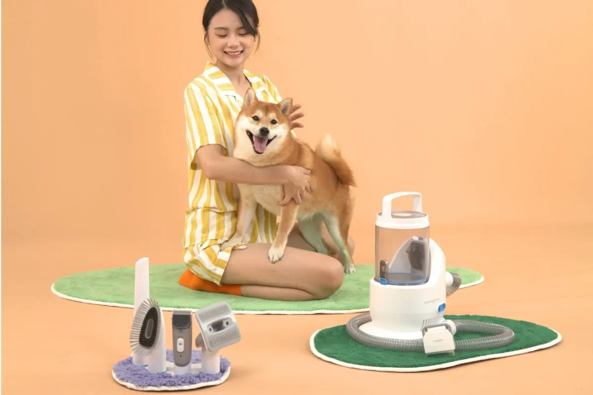 笑顔の女性が柴犬を抱き、ペット用グルーミング製品に囲まれている。犬の毛のケアやトリミングができる電動バリカン、ブラシ、毛吸引器などが並び、ペットとの快適な暮らしを提案する場面。