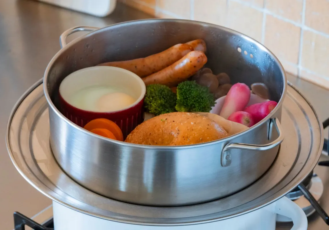 ガスコンロに置かれた蒸し器で、ソーセージ、ブロッコリー、卵、パンなど様々な食材が蒸されている様子