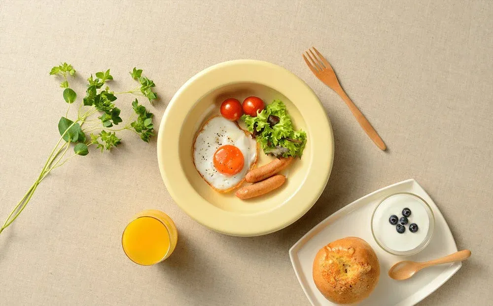 明るい背景に、目玉焼き、ソーセージ、ミニトマト、レタスが盛られたプレートが中央に配置されています。オレンジジュース、ベーグル、ブルーベリーヨーグルトも添えられ、健康的でバランスの取れた朝食の風景です。