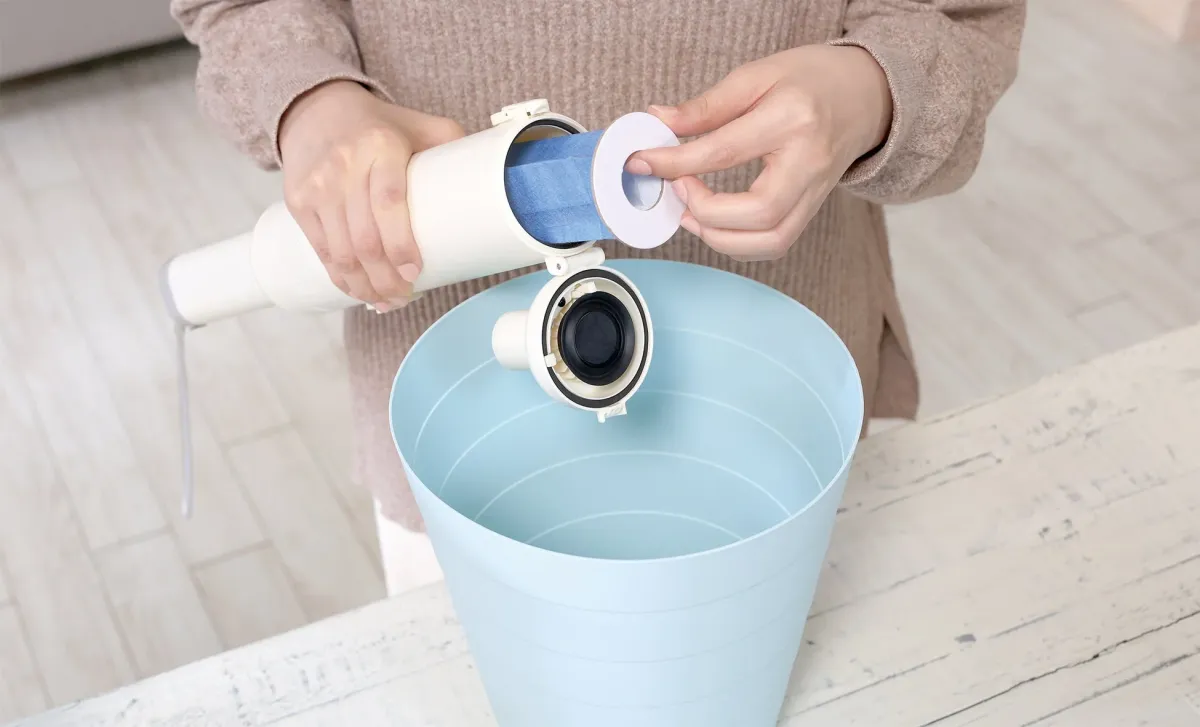 白い筒状の機器から青いフィルターを取り外し、または取り付けようとしている人物の手元を捉えた画像です。水色のバケツが手前にあり、機器のメンテナンスやフィルター交換の様子を示しています。