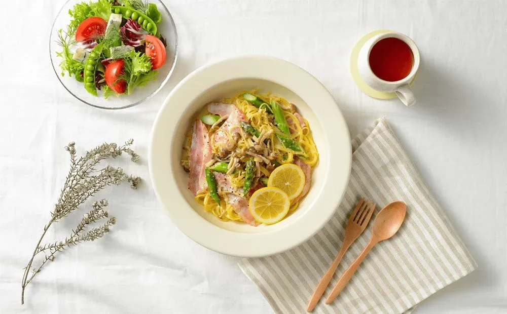 白いテーブルクロスの上に、ベーコンとアスパラガス、きのこ、レモンを添えたクリーミーなパスタ、フレッシュなサラダ、そして紅茶が並べられた食卓の様子。木製カトラリーとストライプ柄のナプキンが添えられ、明るく美味しそうな食事のシーンを捉えています。