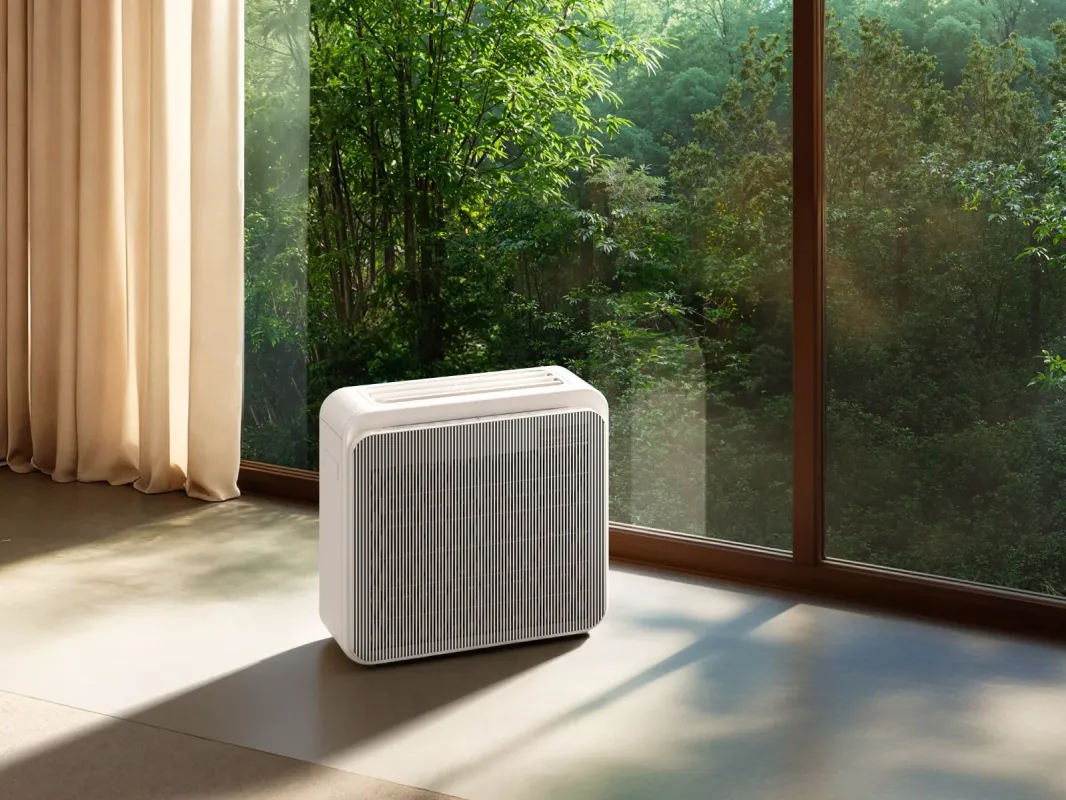 大きな窓から緑豊かな森が見える部屋に、白いモダンな空気清浄機が置かれています。