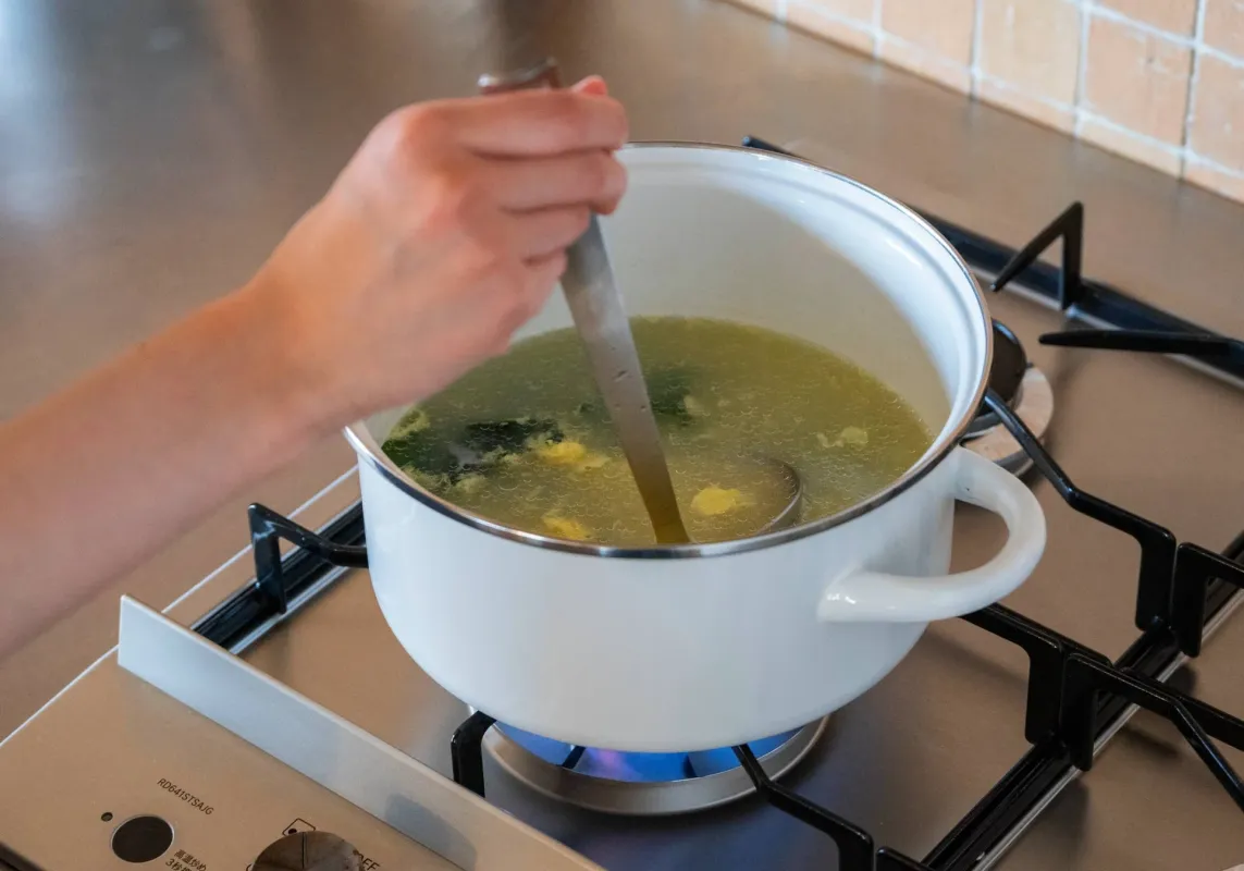 ガスコンロで白い鍋に入ったスープをかき混ぜている様子