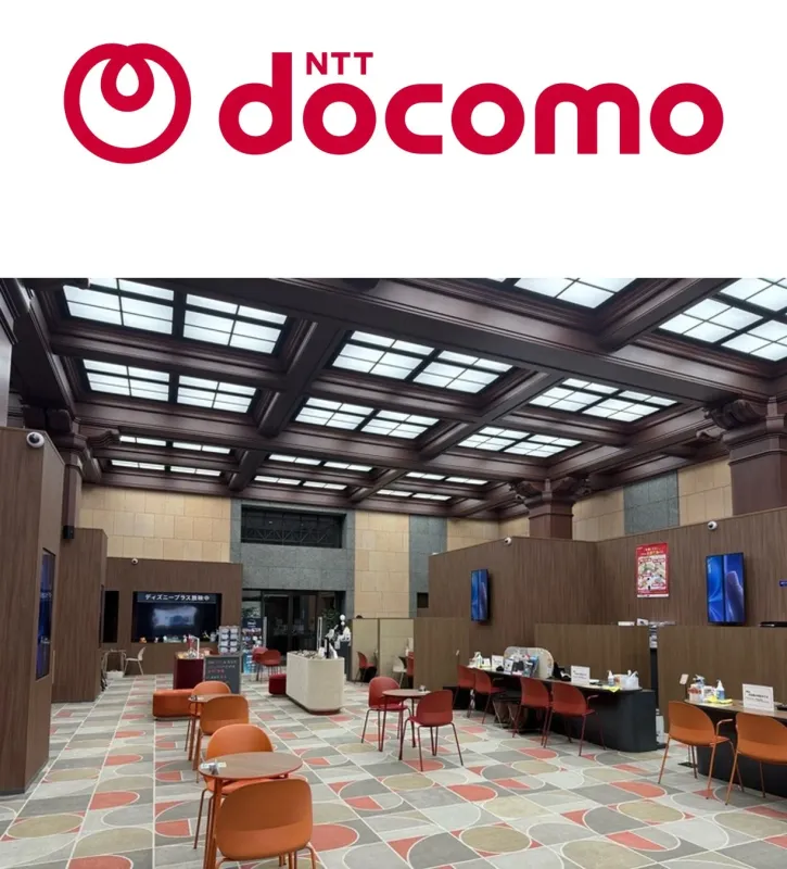 NTTドコモのロゴが上部に表示された、広々とした店舗またはラウンジの内部の様子です。木目調の内装と特徴的な天井、オレンジや赤の椅子が配置された座席エリア、そしてサービスカウンターが見られます。