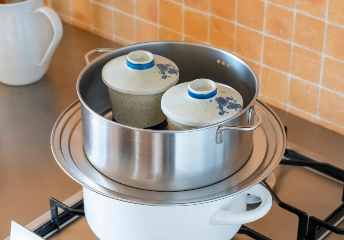 キッチンでガスコンロの上に蒸し器が置かれ、中に2つの蓋付き陶器の器が入っている様子