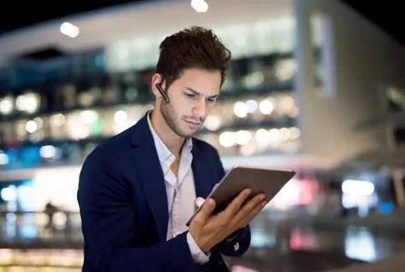 夜の屋外で、スーツを着用した若い男性がBluetoothヘッドセットを装着し、タブレットを真剣な表情で操作しています。