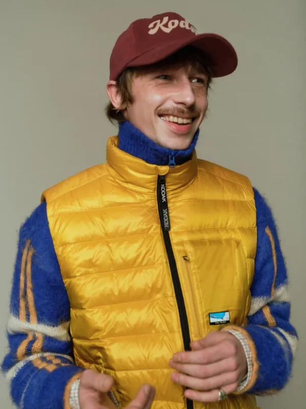 笑顔の若い男性が、黄色いダウンベストと青いモヘアセーター、赤い「Kodak」キャップを着用したカジュアルなファッションポートレート。明るい色彩が特徴的です。