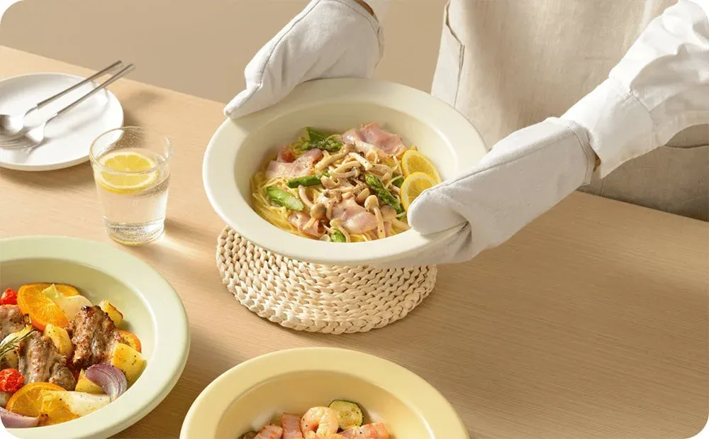 オーブンミトンを着用した人物が、アスパラガス、ベーコン、きのこ、レモンを添えたパスタをテーブルに置いている様子。他にもロースト料理などが並び、食卓が準備されている場面です。