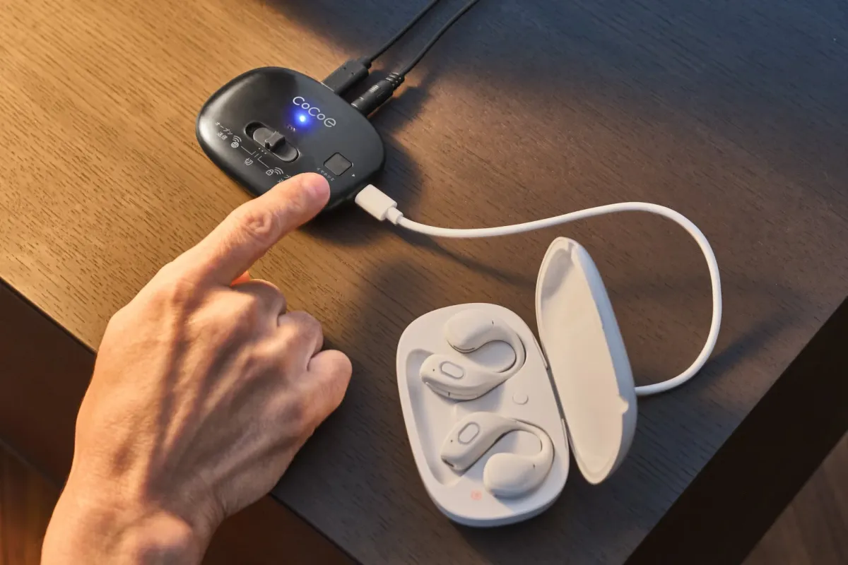 CoCoeと書かれた黒い音響機器を指で操作している様子。隣には充電ケースに入った白いワイヤレスイヤホンがあり、機器とケーブルで繋がっています。