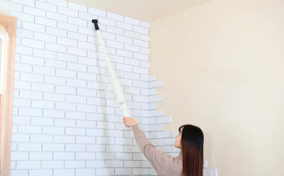 女性が白いレンガ調の壁を棒状の掃除機で掃除している様子です。部屋の清潔感を保つための家事の瞬間を捉えています。