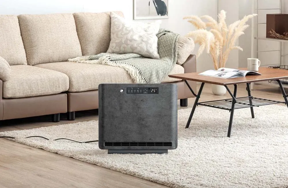 ベージュのソファが置かれたリビングルームに、スタイリッシュなダークグレーの暖房器具が白いふわふわのラグの上に設置されています。木製のコーヒーテーブルには雑誌とマグカップがあり、全体的に落ち着いたモダンな雰囲気です。