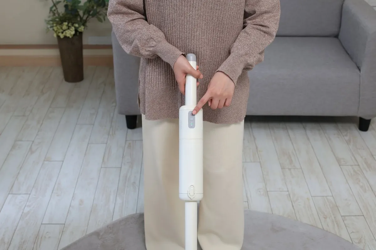 女性がリビングで白いスティック型クリーナーを操作している様子。指が電源ボタンに触れており、バッテリー残量やモードを示すインジケーターが見えます。
