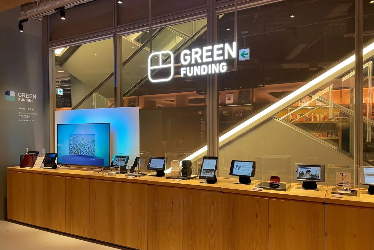 「GREEN FUNDING」のロゴがある店舗の体験型展示スペースで、クラウドファンディング中の未発表新製品が多数ディスプレイされています。来場者が触れて体験できるよう、タブレットや電子機器が展示されており、奥にはエスカレーターが見えます。