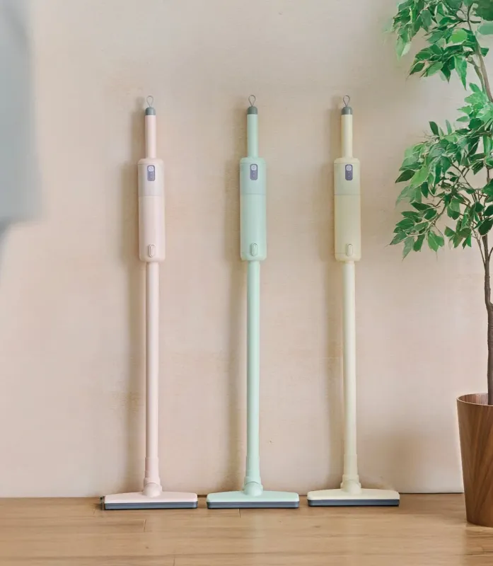 パステルカラーのピンク、ミントグリーン、クリーム色の3本のスティック型フロアクリーナーが、明るい色の壁に立てかけられています。右端には観葉植物が置かれており、清潔感のあるシンプルな空間を演出しています。
