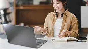 笑顔の若い女性がノートパソコンに向かって話しており、オンラインでのコミュニケーションや学習、リモートワークをしている様子が伺えます。