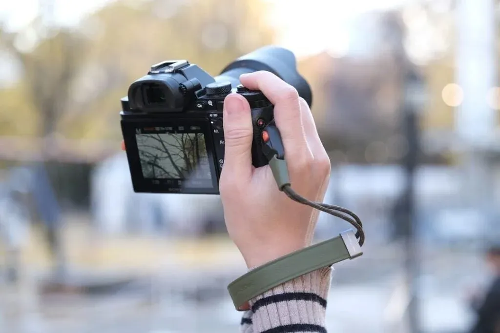 屋外で、カメラの液晶画面に木々が映っている様子を写しながら、手首にリストストラップを装着した手がミラーレスカメラを構えているクローズアップ画像