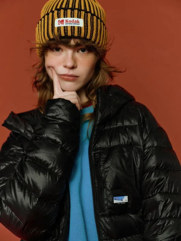 オレンジ色の背景の前でポーズをとる若い女性のポートレート。コダックのロゴが入ったニット帽と黒いダウンジャケットを着用し、顎に手を当てており、カジュアルでファッショナブルな印象です。