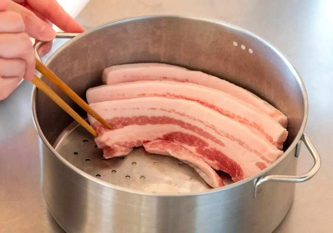 手で箸を使い、生の豚バラ肉の塊を蒸し器に並べている様子