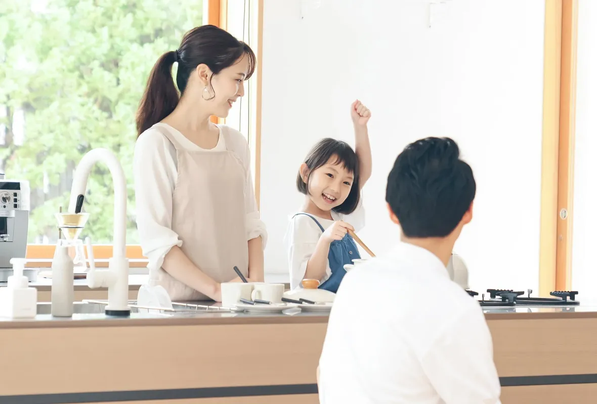 明るいキッチンで、母親と娘が楽しそうに料理をしている家族の風景です。娘は腕を上げて満面の笑みを浮かべ、父親も一緒に過ごしています。温かい家庭の日常が感じられます。
