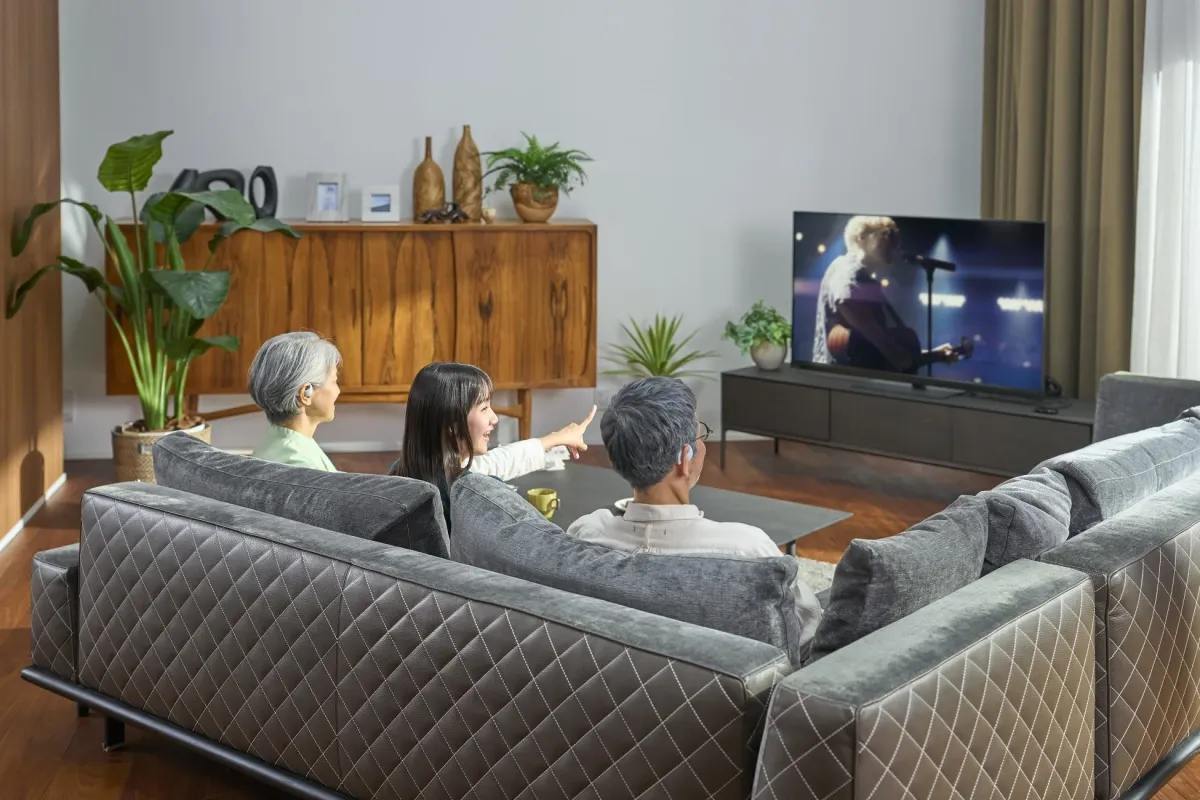 リビングでくつろぐ家族3人がソファに座ってテレビを見ているシーン。若い女性が楽しそうにテレビ画面を指差し、和やかな家族の時間が流れている。