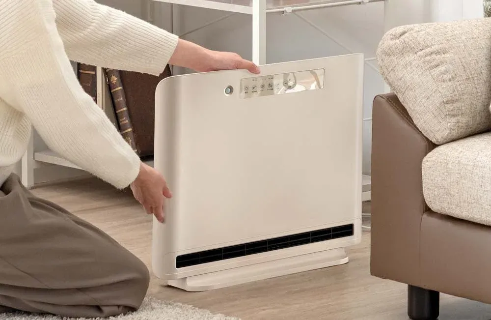 リビングでベージュ色の多機能家電(暖房、送風、加湿など)を床に置いている女性の様子。部屋を快適にするための準備をしている瞬間を捉えた一枚です。