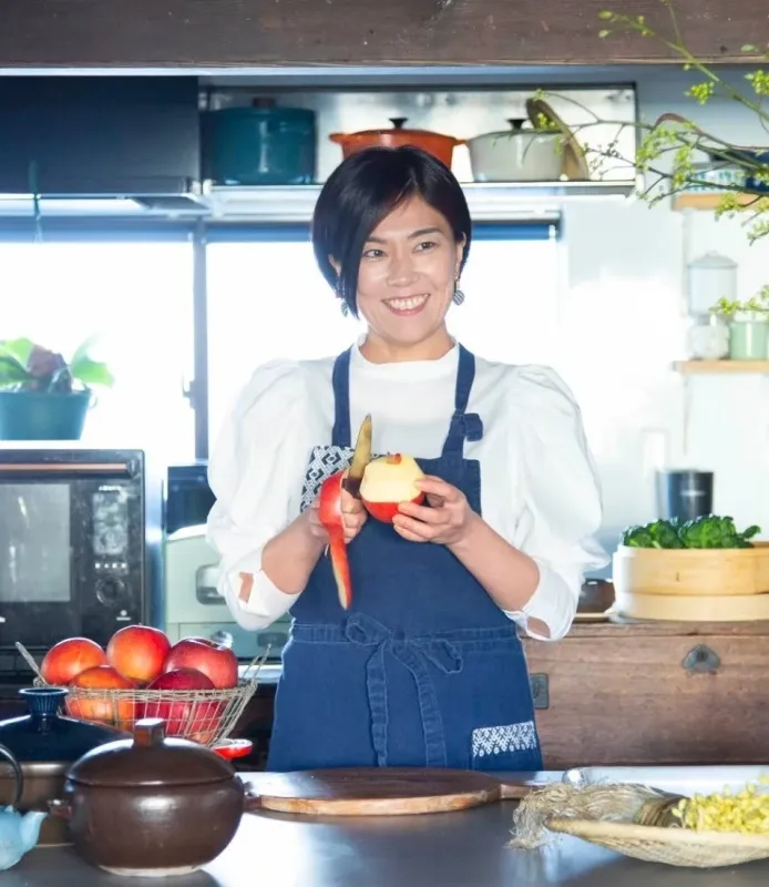 明るいキッチンで、笑顔の女性が青いエプロンを着てリンゴの皮をむいています。