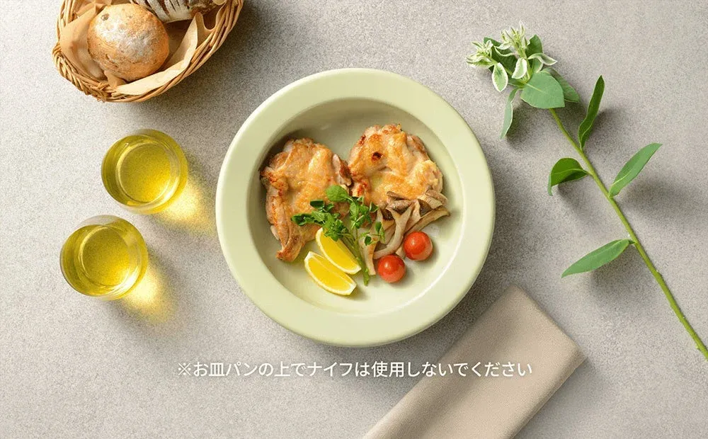 淡いグリーンの皿に盛られたチキンソテーがメインの食卓風景です。付け合わせにレモン、ミニトマト、キノコ、ハーブが添えられ、バスケットにはパン、グラスには飲み物が見えます。全体的に明るく清潔感のある雰囲気です。