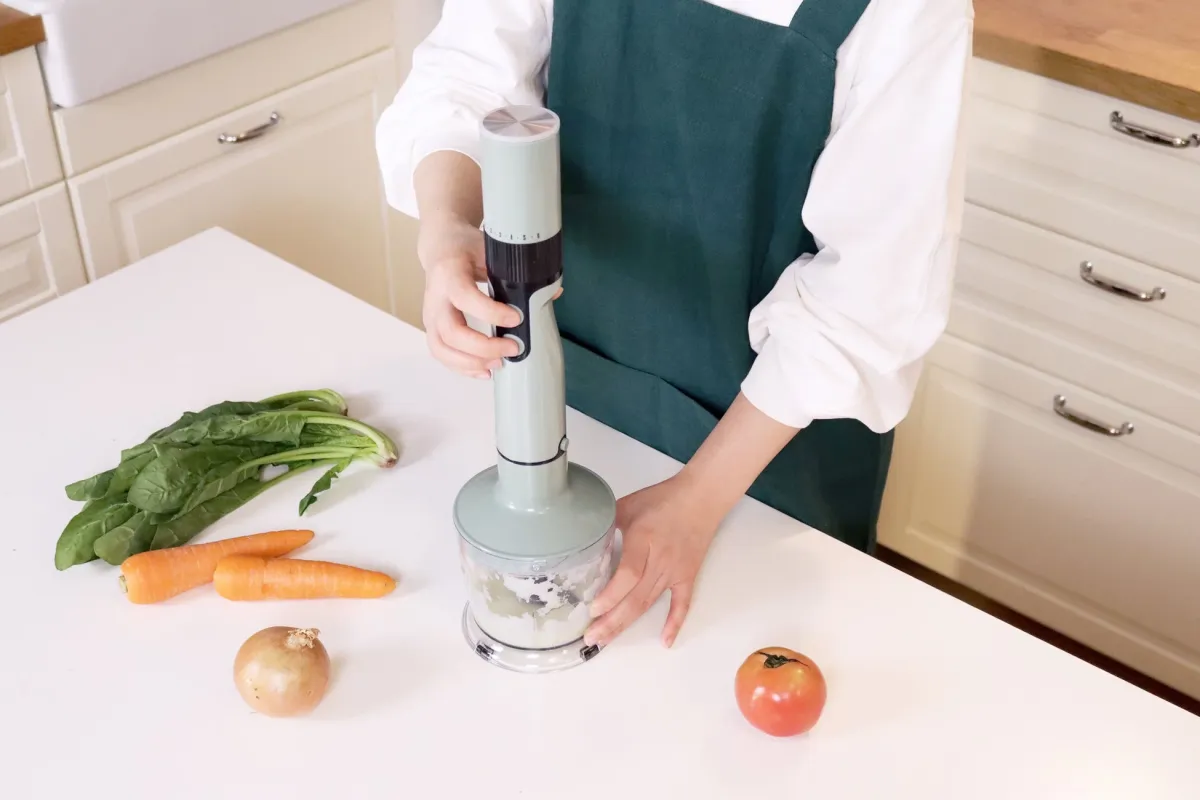 白いキッチンカウンターで、緑色のエプロンをつけた人物がハンドブレンダーを使って野菜を刻んでいます。周りにはほうれん草、人参、玉ねぎ、トマトなどの新鮮な野菜が置かれており、料理の準備をしている様子が伺えます。