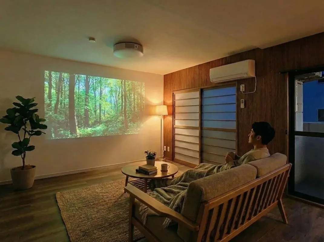 落ち着いたリビングで、男性がソファに座り、壁に投影された森林の映像を眺めてリラックスしている様子。間接照明が温かい雰囲気を作り出しています。