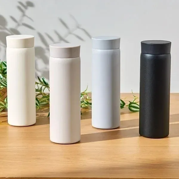 木製のテーブルに、オフホワイト、ベージュ、ライトグレー、ブラックの4色のマットな水筒が並べられています。背景には緑の植物が配置され、ミニマルでモダンな雰囲気を醸し出しています。