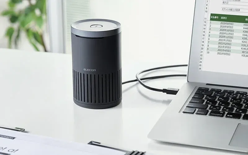 白を基調としたデスクに、ELECOM製の黒い円筒形デバイスがUSBケーブルでノートパソコンに接続されています。パソコン画面にはスプレッドシートが表示され、背景には観葉植物がぼやけて写っています。