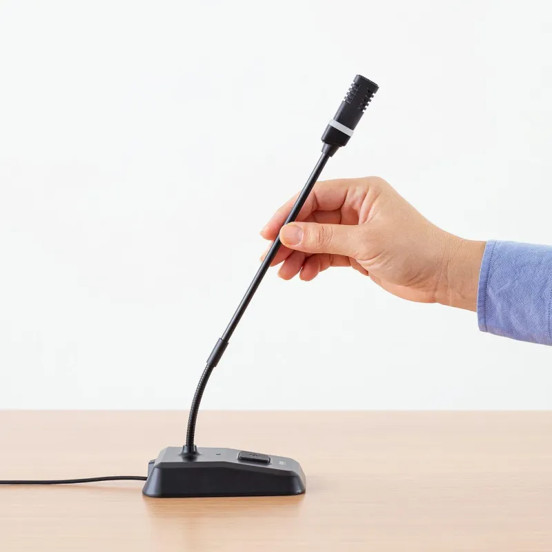 白い背景の木製デスクに置かれた黒いグースネックマイクを、青いシャツを着た人物の手が調整している様子