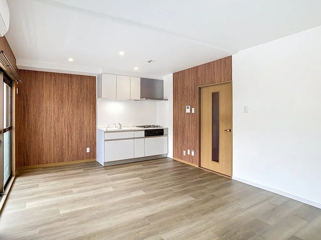 明るい日差しが差し込むモダンなワンルームの室内です。木目調のアクセントウォールと白い壁が特徴で、コンパクトなシステムキッチンが備え付けられています。フローリングの床が広がり、シンプルで清潔感のある空間は、新生活を始めるのに適しています。