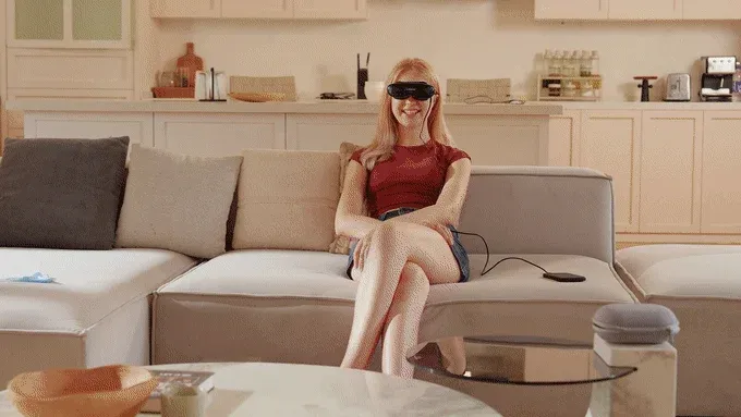 リビングのソファに座り、黒いアイマスクのようなデバイスを装着して笑顔を見せている若い女性の姿。快適な空間でリラックスしている様子がうかがえます。