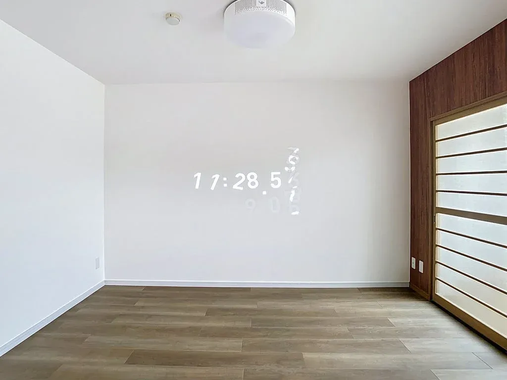 白い壁と木目調のフローリングが特徴のモダンな部屋です。壁にはプロジェクターで「17:28 57」と時刻がデジタル表示されており、右側には障子のような引き戸があります。シンプルで機能的な空間です。