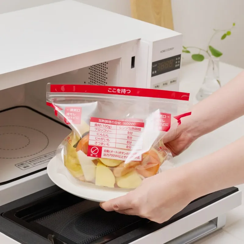 電子レンジで調理する準備をしている様子です。カットされた野菜が入ったクッキングバッグを皿に乗せ、レンジに入れようとしています。バッグには加熱時間や注意事項が書かれており、手軽な蒸し料理の準備を示唆しています。