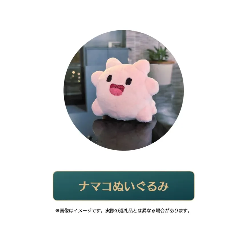 ピンク色で可愛らしいナマコをモチーフにしたぬいぐるみが、室内のぼやけた背景を前に写っています。愛らしい表情が特徴的です。