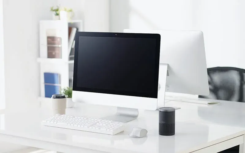 白いデスクに大型モニター、白いキーボードとマウスが整然と並ぶ、ミニマリストでモダンなワークスペースの画像です。タンブラーや小型デバイスも配置され、清潔感のある空間が広がっています。