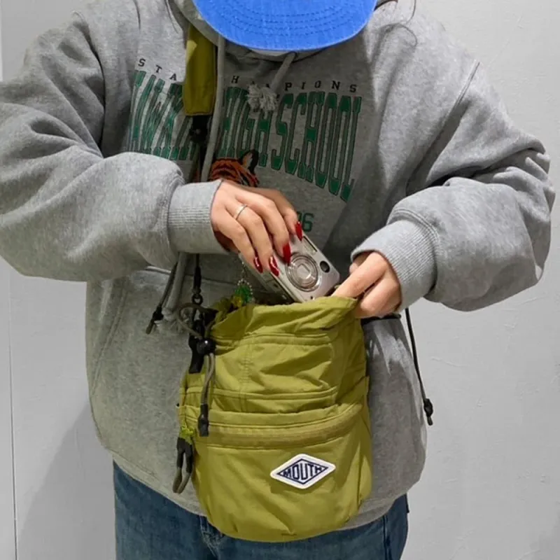 グレーのパーカーとデニムを着用し、青いキャップをかぶった人物が、オリーブグリーンのショルダーバッグにコンパクトカメラをしまっている様子